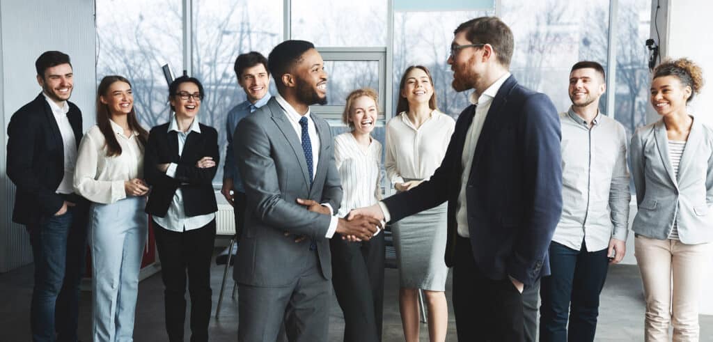 two businessmen handshake while smiling team behind them watch at a New York IT company