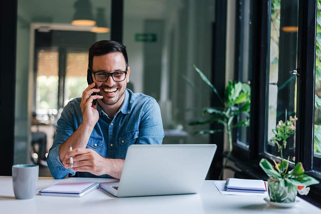 Portrait of young smiling cheerful entrepreneur in casual office making phone call while working with laptop at an IT services company