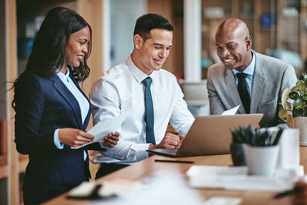 Smiling group of diverse businesspeople going over paperwork together and working on a laptop at a table in an office of an IT company